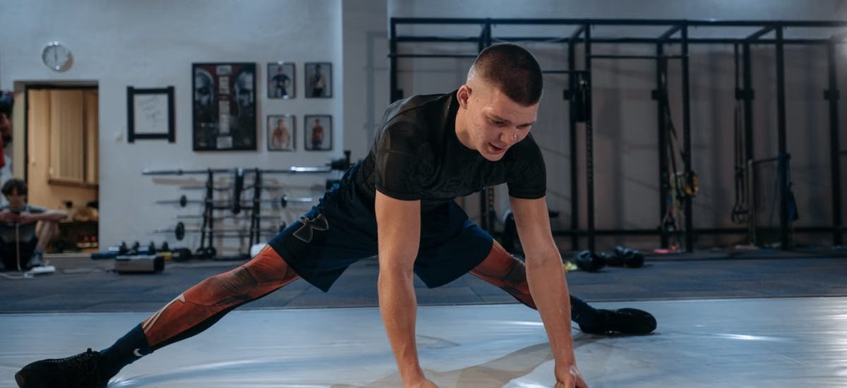 A male athlete stretches intensely in a dimly lit gym environment, preparing for training.